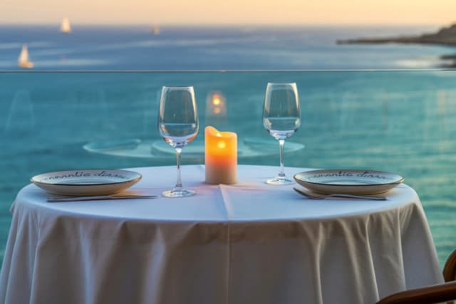 Romantic table for two with wine glasses and a candle on a balcony overlooking the ocean at sunset, with sailboats in the distance.