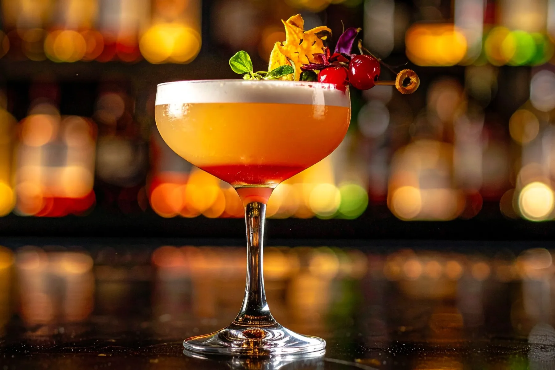 Craft cocktail in a coupe glass with foam and a decorative garnish, set against warm, blurred bar lights.